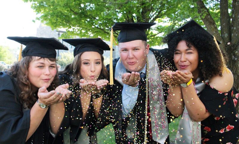 Four 2019 graduates in caps and gowns, blowing glitter from their hands. 